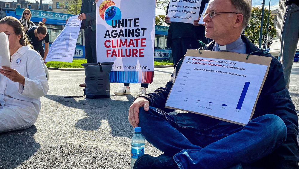 Jörg Alt (r.), Sozialwissenschaftler, Sozialethiker und Migrationssoziologe, sitzt auf dem Boden mit einem Pappschild um den Hals mit einer Grafik mit der Überschrift 'Die Klimakatastrophe macht bis 2070 über 3 Milliarden Menschen zu Geflüchteten', b