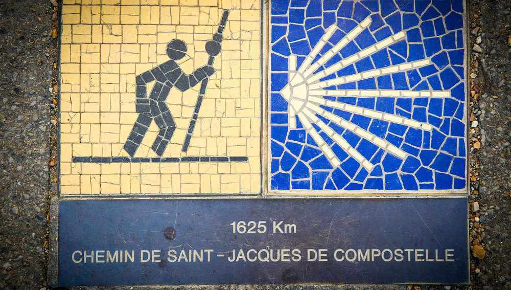 Wegweiser an einer Hauswand mit dem Piktogramm eines Pilgers und gelber Muschel für den Jakobsweg am 15. Juli 2019 in Chartres. Darunter ist die Entfernung nach Santiago de Compostela mit 1625 Km angegeben.